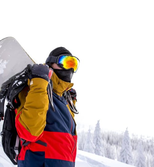 snowboarder-en-gafas-protectoras-sosteniendo-su-tabla-de-snowboard-sobre-su-espalda-scaled-min